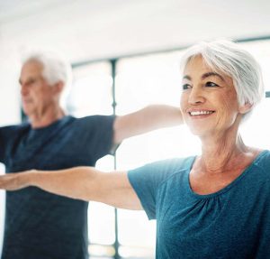 An Elderly Couple Holds Their Arms Outwards From Their Sides In A Yoga Pose.