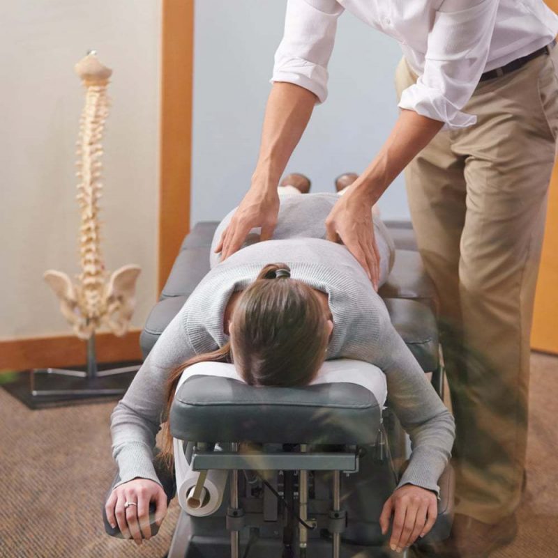 A Female Chiropractic Doctor Stands Over A Female Patient Lying Facedown To Adjust Her Back.