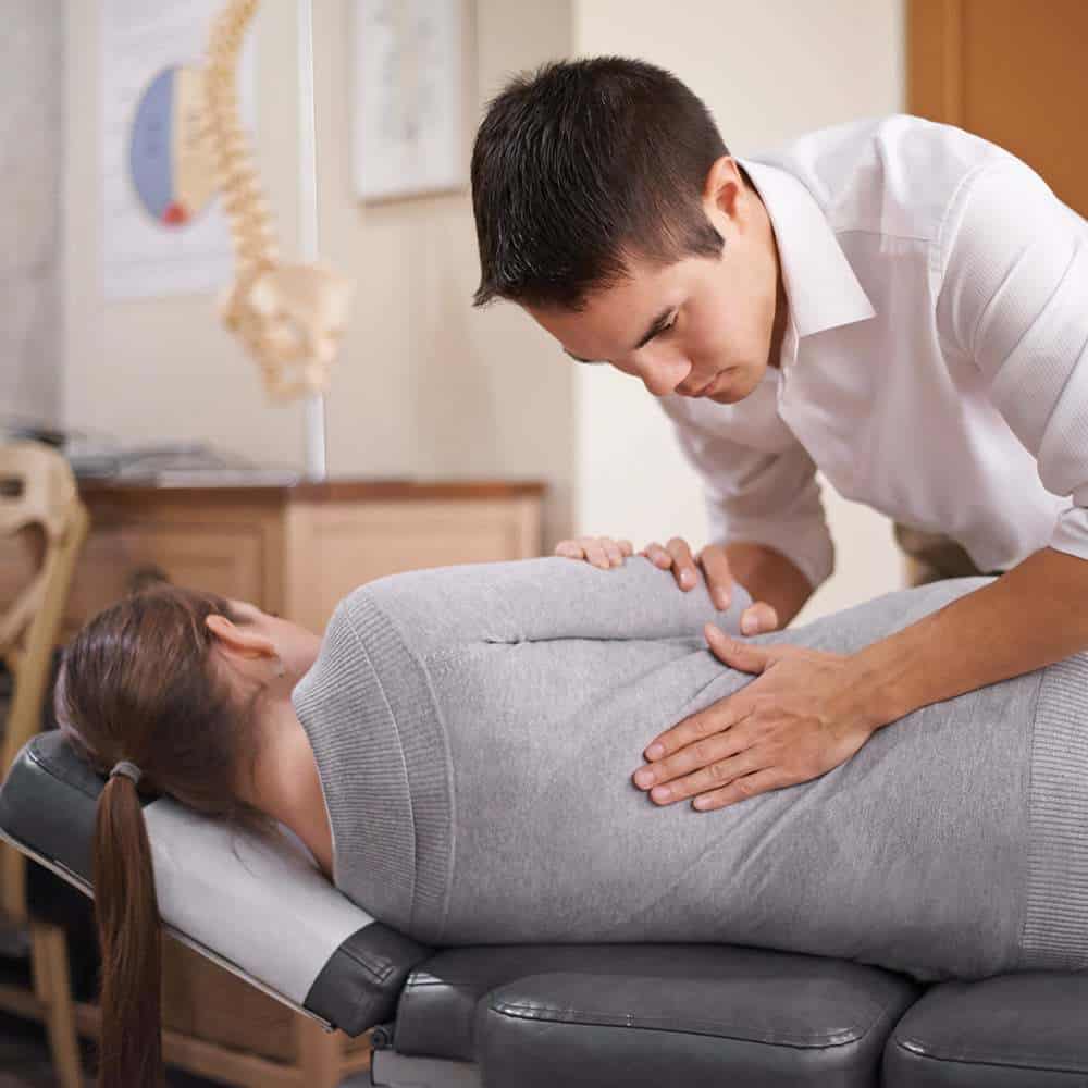 A Male Chiropractic Doctor Stands Over A Female Patient Lying Down To Adjust Her Back.